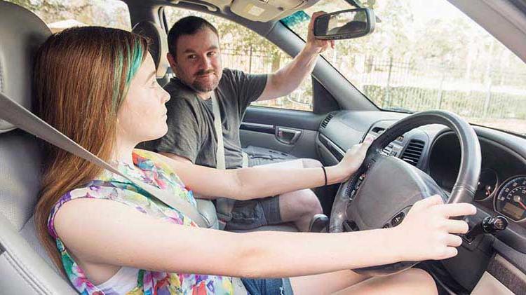 青少年 girl in the driver's seat of a car, buckled up with hands on the wheel. Her father is in the passenger seat adjusting the rearview mirror. 